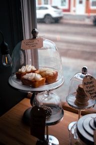 Three lemon ricotta cakes are displayed on a metallic stand under a glass dome. Each cake is topped with creamy frosting. Two additional treats are under separate glass domes: one contains almond flour peanut butter cookies stacked neatly, and another has a chocolate-coated dessert with a tag. The scene is set near a window with raindrops, and the background shows a blurred street with a parked car.