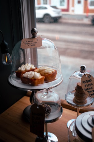 Three lemon ricotta cakes are displayed on a metallic stand under a glass dome. Each cake is topped with creamy frosting. Two additional treats are under separate glass domes: one contains almond flour peanut butter cookies stacked neatly, and another has a chocolate-coated dessert with a tag. The scene is set near a window with raindrops, and the background shows a blurred street with a parked car.