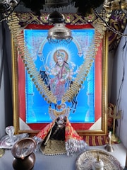A vibrant, ornate image of a deity dressed in colorful garments and adorned with gold jewelry, prominently displayed within a decorative frame. The setting includes religious items such as a trident and oil lamp, suggesting a shrine or altar arrangement.