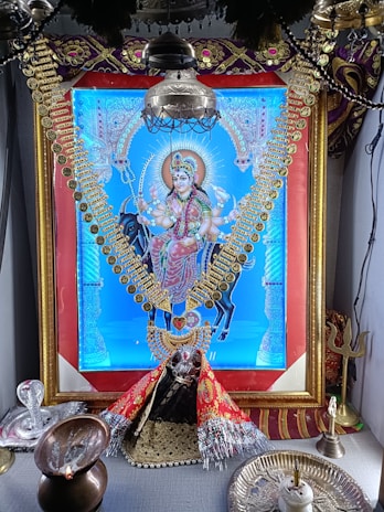 A vibrant, ornate image of a deity dressed in colorful garments and adorned with gold jewelry, prominently displayed within a decorative frame. The setting includes religious items such as a trident and oil lamp, suggesting a shrine or altar arrangement.