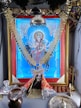 A vibrant, ornate image of a deity dressed in colorful garments and adorned with gold jewelry, prominently displayed within a decorative frame. The setting includes religious items such as a trident and oil lamp, suggesting a shrine or altar arrangement.