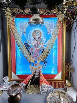 A vibrant, ornate image of a deity dressed in colorful garments and adorned with gold jewelry, prominently displayed within a decorative frame. The setting includes religious items such as a trident and oil lamp, suggesting a shrine or altar arrangement.