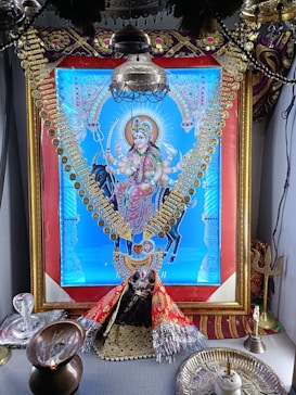 A vibrant, ornate image of a deity dressed in colorful garments and adorned with gold jewelry, prominently displayed within a decorative frame. The setting includes religious items such as a trident and oil lamp, suggesting a shrine or altar arrangement.