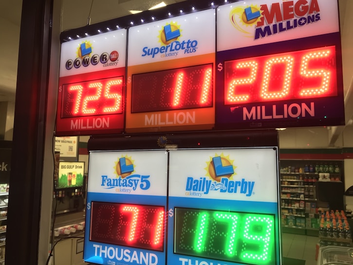 A signboard inside a convenience store displays the current jackpot amounts for various lottery games. There are bright, illuminated numbers in red and green showing the prize amounts: Powerball is at 725 million, Super Lotto Plus at 11 million, Mega Millions at 205 million, Fantasy 5 at 71 thousand, and Daily Derby at 179. In the background, shelves with various drinks and products are visible, along with promotional posters.