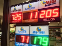 A signboard inside a convenience store displays the current jackpot amounts for various lottery games. There are bright, illuminated numbers in red and green showing the prize amounts: Powerball is at 725 million, Super Lotto Plus at 11 million, Mega Millions at 205 million, Fantasy 5 at 71 thousand, and Daily Derby at 179. In the background, shelves with various drinks and products are visible, along with promotional posters.