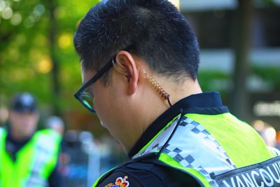 A person wearing a neon yellow reflective vest with a checkered pattern on the shoulder is seen from behind. They have a coiled earpiece in their ear, suggesting a communication device is in use. The background is blurred, with hints of greenery and other individuals.