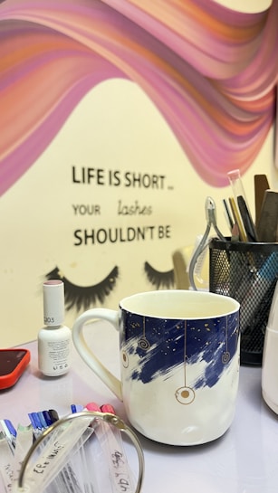 A tabletop with various items including a decorative mug with a blue and white celestial design, a small bottle of nail polish, colorful nail samples, and a metal pen holder with tools. The background features a stylized mural with flowing pink and purple waves and the words 'Life is short ... your lashes shouldn't be' along with painted eyelashes.