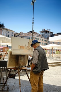 A person in casual attire, wearing a hat, paints an outdoor scene at an easel in a sunlit town square. Surrounding buildings with tiled roofs and shutters frame the scene, while large umbrellas provide shade to nearby seating areas.
