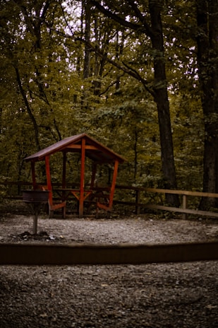 A small wooden gazebo with a red roof and simple bench seating sits on a gravel area surrounded by a wooden fence. It is nestled within a dense forest characterized by tall trees and abundant foliage. The scene is in a natural, outdoor setting.
