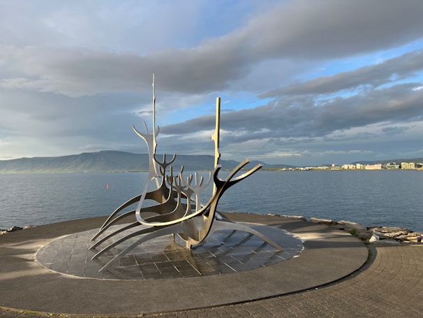 A striking metal sculpture resembling a Viking ship stands on a paved area by the sea. The sky is partly cloudy, with a mountain range visible across the water in the background. On the horizon, city buildings can be seen, basking in soft light.