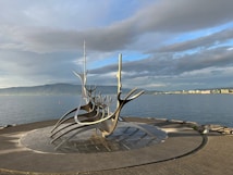A striking metal sculpture resembling a Viking ship stands on a paved area by the sea. The sky is partly cloudy, with a mountain range visible across the water in the background. On the horizon, city buildings can be seen, basking in soft light.
