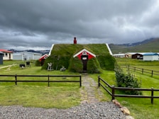 A small house with a grass-covered roof is situated in a rural landscape. It features red and white accents and is surrounded by a lawn with various garden tools and objects. Several similar houses can be seen in the background under a cloudy sky, with hilly terrain in the distance.