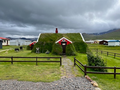A small house with a grass-covered roof is situated in a rural landscape. It features red and white accents and is surrounded by a lawn with various garden tools and objects. Several similar houses can be seen in the background under a cloudy sky, with hilly terrain in the distance.