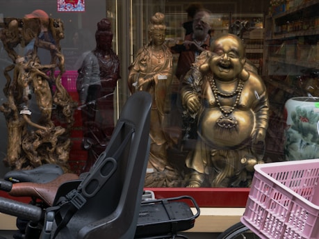 A store window displays a variety of items including ornate wooden sculptures, a golden laughing Buddha statue, and another smaller figure. In the foreground, a bicycle with a child seat and a pink basket is parked in front. The reflection of the street and pedestrians can be seen on the glass.