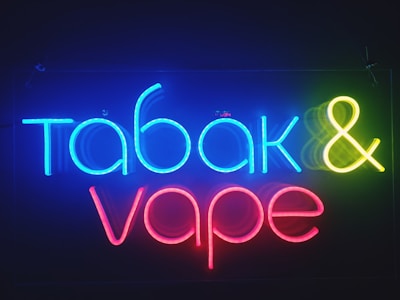 Bright neon sign displaying the words 'tabak & vape' in colorful letters against a dark background. The word 'tabak' is in blue, the ampersand is in yellow, and 'vape' is in red.
