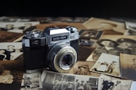 A vintage Zeiss Ikon camera is prominently placed among a scattered collection of old sepia-toned photographs. The camera's metallic body and lens give it a classic, historical look, surrounded by images displaying various portraits and family scenes.