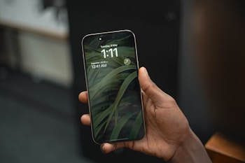 A hand holds a smartphone displaying the lock screen showing the time as 1:11 on Tuesday, 9 May, with a background of green leaves.