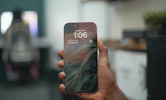 A person is holding a smartphone in their hand. The phone displays the time as 1:06 on a Tuesday, 9 May. The lock screen features an artistic wallpaper with leafy patterns. The background is slightly blurred, indicating an indoor setting with another figure seated at a desk.
