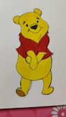 A yellow cartoon bear with a red shirt stands with a cheerful expression. The character has small ears, black eyes, a nose, and a friendly smile. The background is white with a partial view of a red and pink floral pattern at the bottom edge.