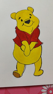 A yellow cartoon bear with a red shirt stands with a cheerful expression. The character has small ears, black eyes, a nose, and a friendly smile. The background is white with a partial view of a red and pink floral pattern at the bottom edge.