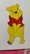 A yellow cartoon bear with a red shirt stands with a cheerful expression. The character has small ears, black eyes, a nose, and a friendly smile. The background is white with a partial view of a red and pink floral pattern at the bottom edge.