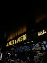 A dimly lit restaurant interior features a neon sign reading 'LA POLLO & PASTA' above a counter. Below the sign, illuminated menu boards display various food options. Two staff members are working behind the counter.