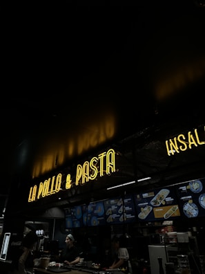 A dimly lit restaurant interior features a neon sign reading 'LA POLLO & PASTA' above a counter. Below the sign, illuminated menu boards display various food options. Two staff members are working behind the counter.