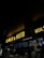 A dimly lit restaurant interior features a neon sign reading 'LA POLLO & PASTA' above a counter. Below the sign, illuminated menu boards display various food options. Two staff members are working behind the counter.