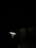 A single streetlight illuminates a small area in an otherwise dark setting. The light emits a bright, focused beam that highlights the pole and a small part of the surroundings. There are shadows cast on adjacent structures, hinting at an urban environment.