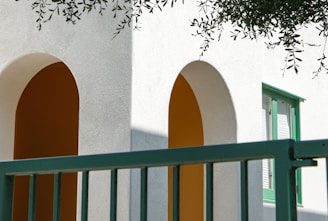 A minimalist architectural design featuring white walls with orange arches casting shadows. The structure is accented by green window frames and a matching green railing in the foreground. Olive branches hang down from the top of the image, adding a natural element.