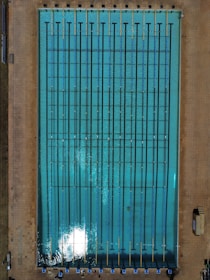 An aerial view of an Olympic-sized swimming pool with clear blue water, marked with dark lane dividers and surrounded by a tiled deck. Pool chairs and some equipment are visible along the lower edge, while shadows suggest sunlight on the water's surface.