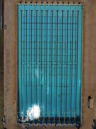 An aerial view of an Olympic-sized swimming pool with clear blue water, marked with dark lane dividers and surrounded by a tiled deck. Pool chairs and some equipment are visible along the lower edge, while shadows suggest sunlight on the water's surface.