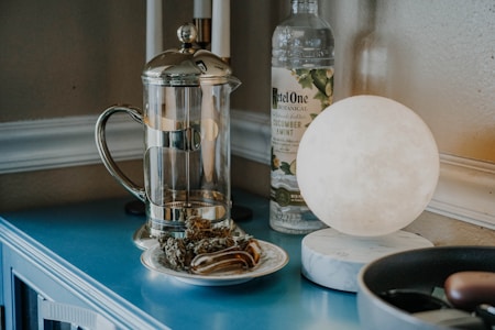A kitchen countertop setup featuring a glass and metal French press, a bottle of cucumber mint infuser, a glowing round lamp, and a small plate of dried herbs or tea leaves. The surface is a bright blue color, which contrasts with the neutral tones of the objects.