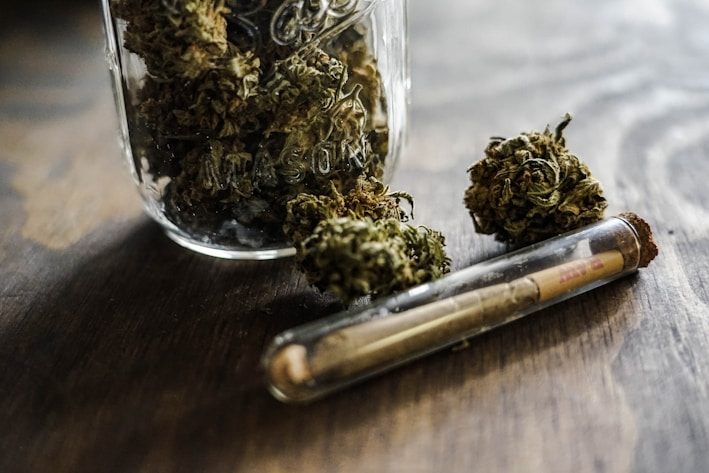 A glass mason jar filled with dried cannabis buds on a wooden surface, accompanied by a pre-rolled joint inside a glass tube.