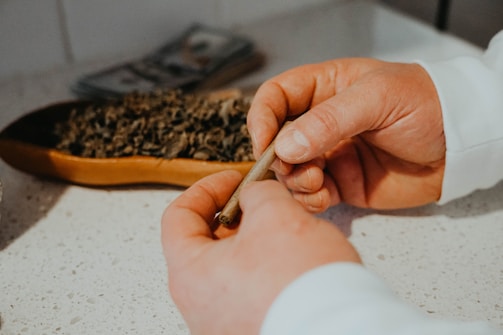 A person is rolling a joint with their hands on a speckled countertop. A wooden tray containing loose tobacco or similar substance is visible nearby.