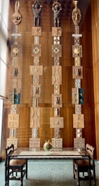 A large wall display featuring three vertical columns of intricate wooden panels and sculptures. Each column has a series of geometric and abstract patterns, with statues and tribal masks at the top. The panels feature earthy tones and complex designs that suggest cultural art. Below the display is a glass table with a floral centerpiece, surrounded by four wooden chairs on a polished stone floor.