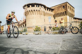 A cyclist in athletic gear rides a bicycle past a historical brick fortress with defensive towers. Another person is cycling in the background, while a spectator in a yellow shirt watches nearby. The area is cordoned off with metal railings, and flags decorate the fortress walls.