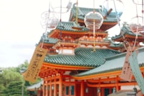 Intricate traditional Japanese architecture is seen in the background, characterized by green-tiled roofs and vibrant orange wooden structures. In the foreground, delicate glass wind chimes and paper tags with Japanese writing hang, gently swaying in the breeze.