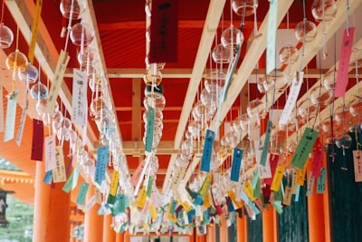 Multiple glass wind chimes are hanging in rows from a wooden structure, each accompanied by colorful strips of paper with writing on them. The setting appears to be a traditional or cultural site, reflected in the vibrant orange columns and beams in the background.