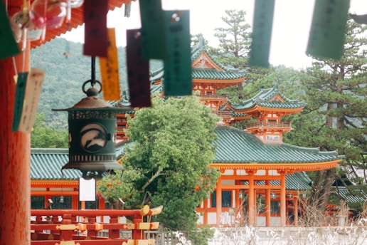 Traditional Japanese architecture with green-tiled roofs and red-orange wooden structures surrounded by lush greenery and trees. A decorative lantern and colorful strips of paper hang prominently in the foreground.