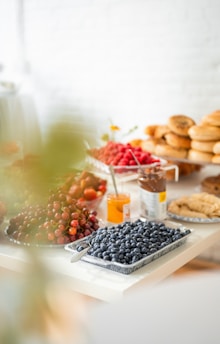A table is set with various breakfast or brunch items, including a plate of blueberries, a bowl of red grapes, sliced bread, a jar of spread, a tray of raspberries, and a stack of bagels. The background is softly blurred, suggesting a bright, airy kitchen or dining area.