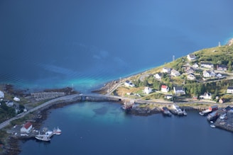 An aerial view of a coastal village with several houses and buildings. The village is situated near a body of clear blue water and features a road that connects to a small bridge over the water. There are a few boats docked at small piers, and the area is lush with greenery and open land.