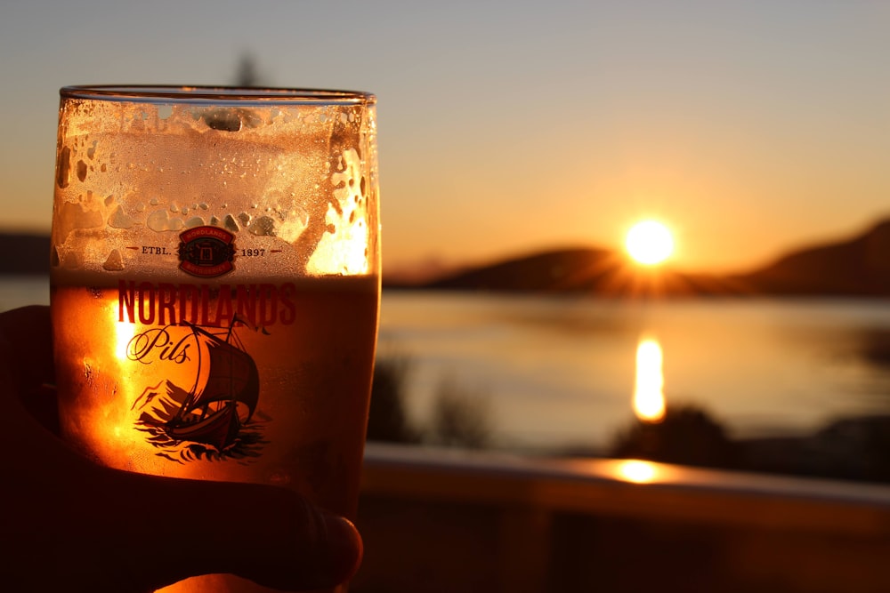 a person holding a glass of beer in front of a sunset