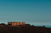 A wooden deck with railings sits on a barren, rocky landscape. The deck is elevated slightly above the ground and is flanked by some construction cones or markers. The sky above is clear with a gradient from light blue to darker shades, suggesting an open and expansive view.