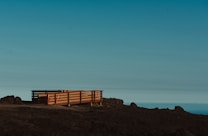A wooden deck with railings sits on a barren, rocky landscape. The deck is elevated slightly above the ground and is flanked by some construction cones or markers. The sky above is clear with a gradient from light blue to darker shades, suggesting an open and expansive view.