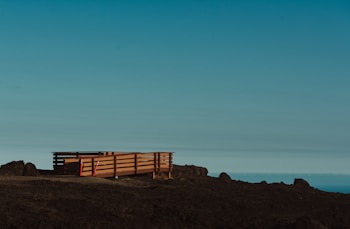 A wooden deck with railings sits on a barren, rocky landscape. The deck is elevated slightly above the ground and is flanked by some construction cones or markers. The sky above is clear with a gradient from light blue to darker shades, suggesting an open and expansive view.