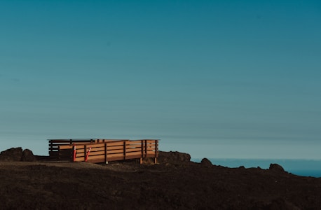A wooden deck with railings sits on a barren, rocky landscape. The deck is elevated slightly above the ground and is flanked by some construction cones or markers. The sky above is clear with a gradient from light blue to darker shades, suggesting an open and expansive view.