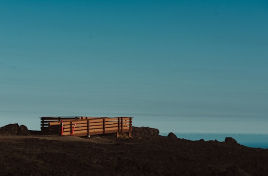 A wooden deck with railings sits on a barren, rocky landscape. The deck is elevated slightly above the ground and is flanked by some construction cones or markers. The sky above is clear with a gradient from light blue to darker shades, suggesting an open and expansive view.