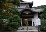 A traditional Japanese building with intricate wooden architecture and a tiled roof, surrounded by lush green trees. The entrance features a small flight of stairs leading to wooden doors and signposts with Japanese inscriptions.