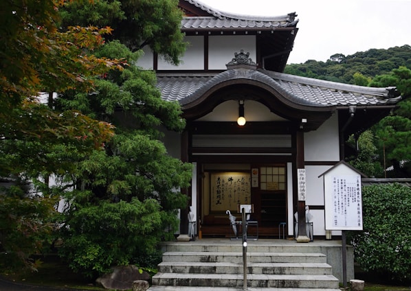 A traditional Japanese building with intricate wooden architecture and a tiled roof, surrounded by lush green trees. The entrance features a small flight of stairs leading to wooden doors and signposts with Japanese inscriptions.